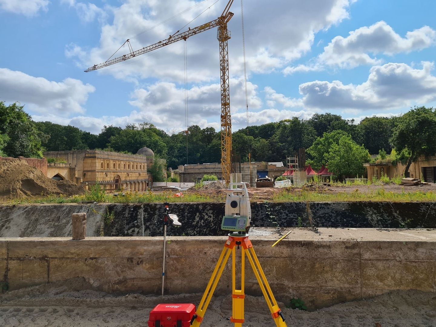 Vermessungsgerät auf gelbem Stativ vor Baustelle mit Kran und historischen Gebäuden im Zoo Hannover bei bewölktem Himmel.