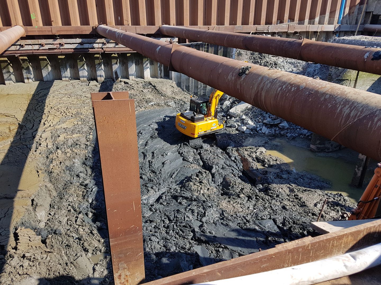 Baustelle mit schlammigem Boden, Stahlträgern und kleinem gelbem Bagger in einer Baugrube mit Stahlverbau.