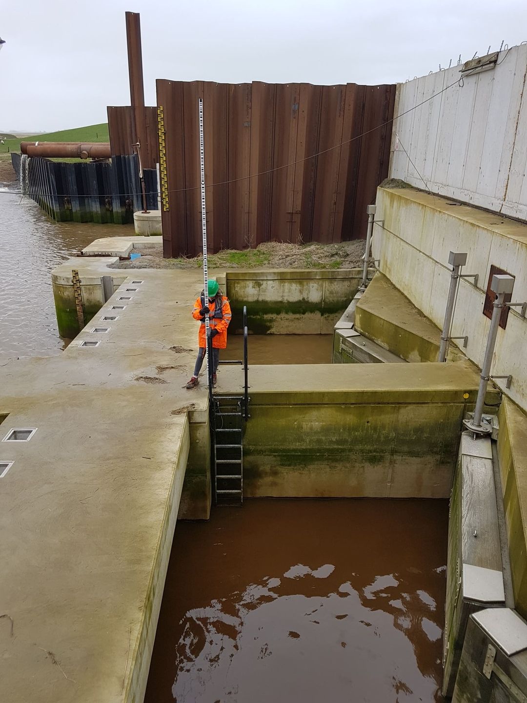 Vermessungstechniker in orangefarbener Schutzkleidung und grünem Helm steht auf einer Leiter an einem Wasserbauwerk und hält eine Messlatte.