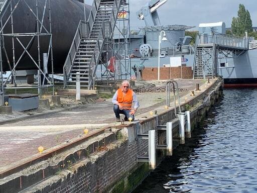 Ein Mann in orangefarbener Arbeitsjacke kniet an einem Hafen