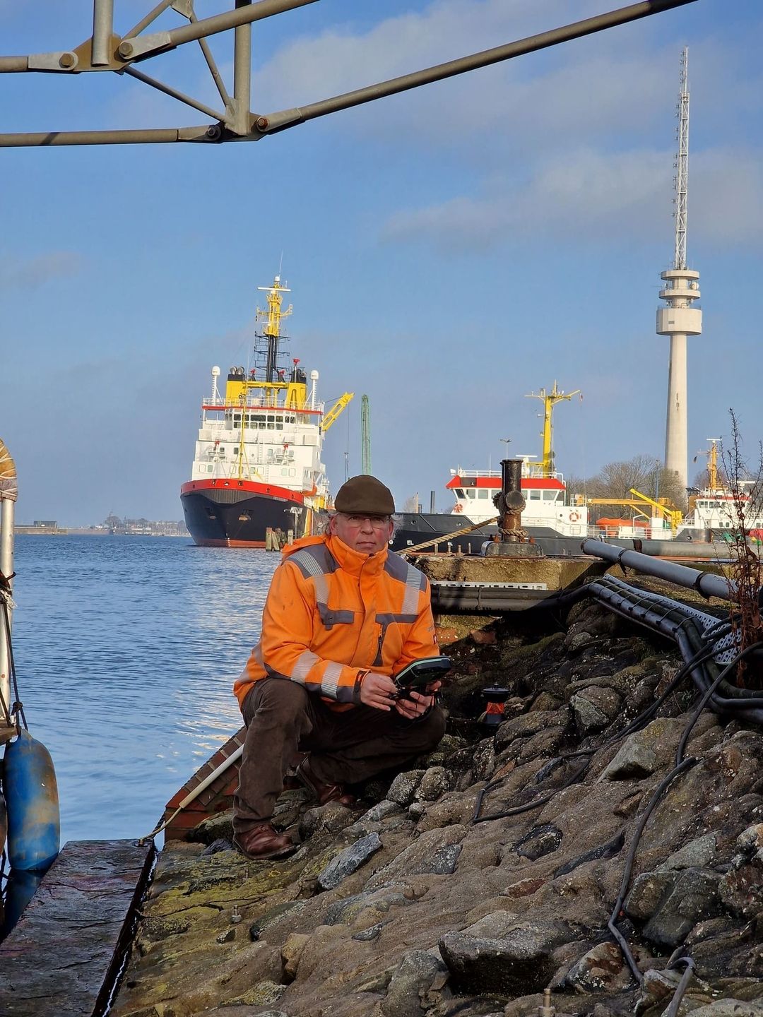 Herr Scholz in orangefarbener Arbeitsjacke sitzt auf steinigem Ufer, hält Gerät in der Hand, im Hintergrund Schiffe und Fernsehturm bei klarem Himmel.