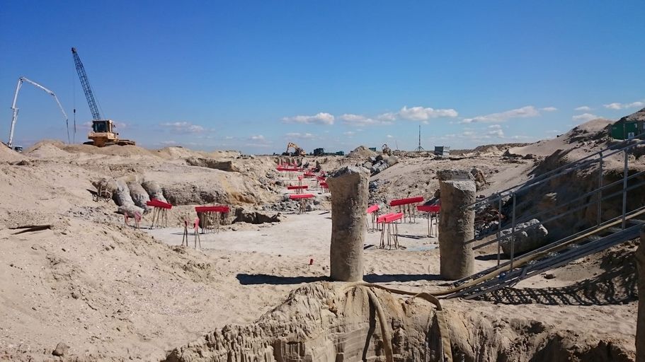 Baustelle mit Erdarbeiten, mehreren Betonpfeilern und rot markierten Messpunkten im Sand, Kran und Bagger im Hintergrund unter blauem Himmel.