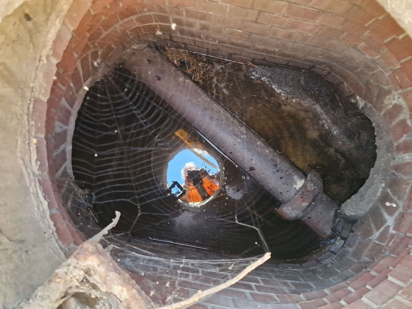 Blick in einen runden Schacht mit Ziegelmauerwerk, darin ein Metallrohr und ein großes Spinnennetz, Spiegelung einer Person in orangefarbener Kleidung im Wasser.